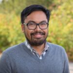 A photo of a filipino man with close cut hair, glasses, and beard. He is wearing a plaid button up under a grey sweater.